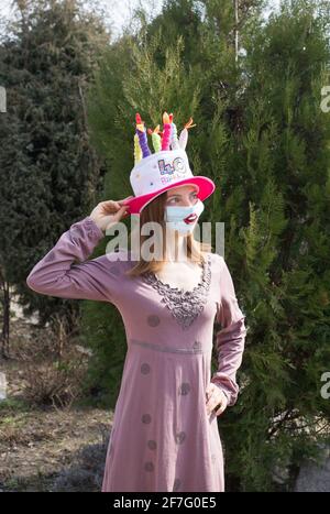 b-Tages-Feier während der Selbstisolation. Geburtstagsfrau in einer medizinischen Maske mit bemalten Lippen, in lustigen Hut in Form von Kuchen mit Kerzen. Humor Konzept, Stockfoto