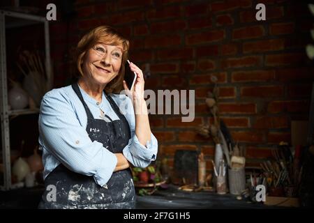 Fröhliche ältere Frau, die in der Töpferwerkstatt am Handy spricht Stockfoto