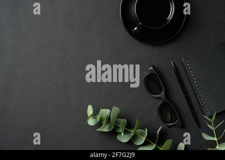 Arbeitsplatz aus schwarzem Leder mit Bürobedarf, Kaffeetasse, Notizbuch aus Papier, Brille, Eukalyptusblättern. Draufsicht mit Kopierplatz, flach liegend. Stockfoto