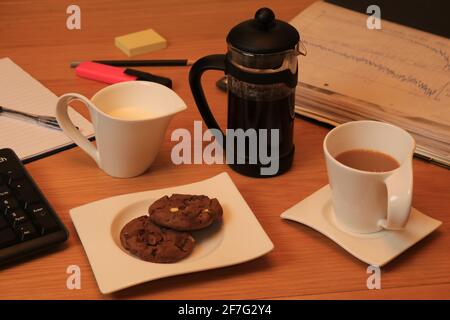 Kaffeetasse und Krug mit Milch, machen Sie eine Pause, trinken Sie und zwei Schokoladenkekse auf dem Teller, Dokumente, Tastatur, Stift, Stift und Pad Stockfoto