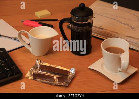 Kaffeetasse und Kanne mit Milch, machen Sie eine Pause, trinken und mit Wafer-Schokolade überzogene Snacks in Zinnfolie, Dokumenten, Tastatur, Stift, Bleistift und Pad Stockfoto