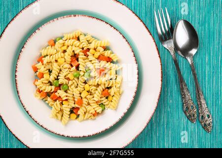 Köstliche, vegetarische Fusilli-Pasta mit Basilikumpesto, Karotten, süßem Mais und grüner Erbse auf einem Teller auf einer türkisfarbenen Tischdecke Stockfoto