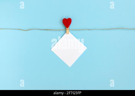 Weißes Stück Papier mit Wäscheklammer am Seil befestigt, auf blauem Hintergrund, Rahmen für glücklichen Valentinstag, leer mit leerem Platz für Text Stockfoto