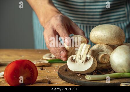 Koch in einer blau gestreiften Schürze hält einen frischen Pilz in der Hand über einem Holztisch mit Gemüse Und andere Zutaten zum Kochen des Gerichts Stockfoto