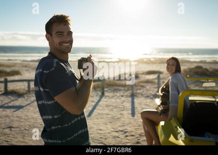 Kaukasisches Paar am Strand, Mann mit Kamera lächelnd, Frau auf Strandbuggy sitzend, Gitarre spielend Stockfoto