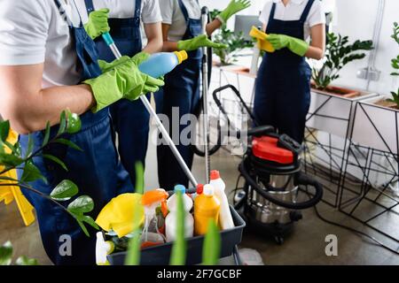 Abgeschnittene Ansicht des Reinigungsmittels in Gummihandschuhen, die das Reinigungsmittel in der Nähe halten Kollegen und Staubsauger im Büro Stockfoto