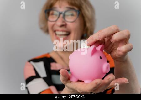 Glückliche ältere Frau legt Münze in Sparschwein auf weißem Hintergrund. Stockfoto