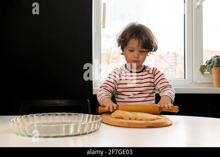 Kleines Kind bereitet einen Kuchen vor und rollt den Teig mit einem Nudelholz auf dem Küchentisch drinnen aus. Stockfoto