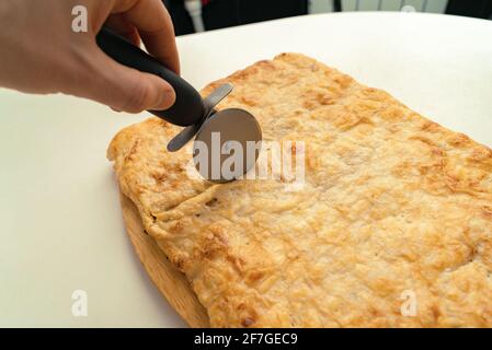 Menschliche Hand schneidet den fertig gebackenen Kuchen mit einem runden Messer auf dem Küchentisch. Stockfoto