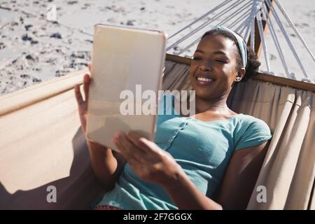 Glückliche afroamerikanische Frau, die in der Hängematte am Strand liegend benutzt Tablet Stockfoto