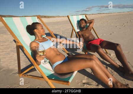 afroamerikanisches Paar in Liebe sitzt auf Liegestühlen und hält sich die Hände am Strand Stockfoto