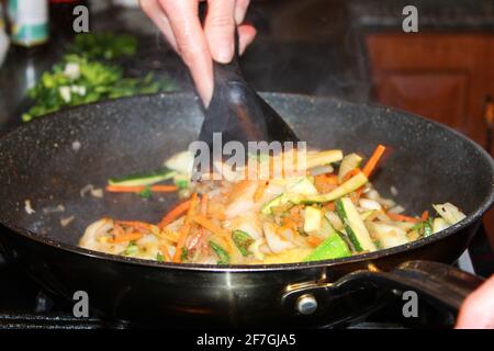 Eine Nahaufnahme einer Person rührt Gemüse an. Teilansicht der Hand auf dem Utensil und der anderen Hand auf dem Griff der Bratpfanne. Stockfoto