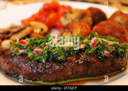 Nahaufnahme eines gegrillten Steak mit Chimichurri-Sauce. Kartoffelkroketten, Pilze und gerösteter roter Pfeffer im Hintergrund unscharf. Stockfoto