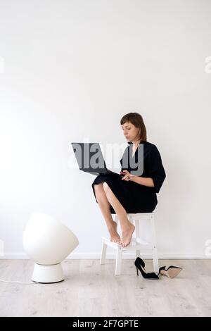 Frau in einem Kleid und barfuß sitzt auf einem Stuhl Mit einem Laptop und funktioniert Stockfoto