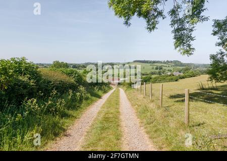 Badby, Northamptonshire, Großbritannien - 28. Mai 2020: Ein unbefestigter Weg zwischen einer Hecke und einem Zaun führt einen Hügel hinunter zu einigen landwirtschaftlichen Gebäuden. Stockfoto