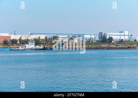 Schubboot schiebt trockenen Massengutfrachter auf dem Fluss Vor dem Hintergrund einer Industrielandschaft am Ufer Stockfoto