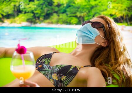 Junge Frau im Badeanzug und Schutzmaske, die sich am Strand mit einem Cocktail entspannt. Exotische tropische Landschaft mit türkisfarbenem Wasser und Palmen. Pandemietourismus Stockfoto