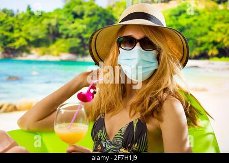 Junge Frau im Badeanzug und Schutzmaske, die sich am Strand mit einem Cocktail entspannt. Exotische tropische Landschaft mit türkisfarbenem Wasser und Palmen. Pandemietourismus Stockfoto