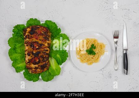 Ein großes Stück Schweinefleisch, im Ofen gekocht, im Ofen gebacken, auf grünen Salatblättern. Mit Pasta auf einem Teller mit einer Gabel und einem Messer. Stockfoto