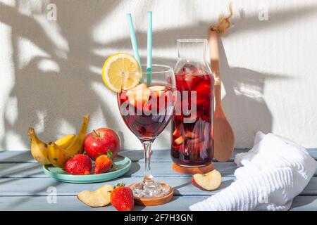 Stillleben eines Glases und einer Flasche Sangria mit einem Obstteller und einem Holzlöffel auf einem sonnigen weißen Hintergrund mit dem Schatten einer Palme. Stockfoto