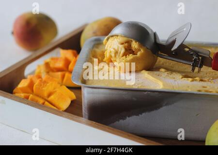 Ausschaufeln von Mango-Eis zum Servieren aus einem Tablett, das außerhalb des Gefrierschranks aufbewahrt wird. Ein Genuss in der Sommerhitze. Stockfoto