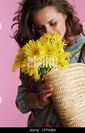 Lächelnde Frau mittleren Alters im Blumenkleid mit gelben Chrysanthemen Blumen und Strohsack auf rosa Hintergrund. Stockfoto