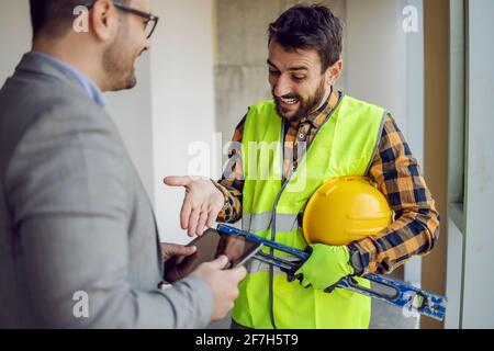 Lächelnder Bauarbeiter zeigt auf die Dokumentation auf dem Tablet und spricht mit seinem Vorgesetzten. Stockfoto