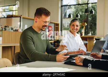 Mann, der am Tisch sitzt und dem Team mit einem digitalen Tablet die neue Strategie erklärt. Kollegen, die eine Strategie planen, sitzen in einem Meeting am Tisch. Stockfoto