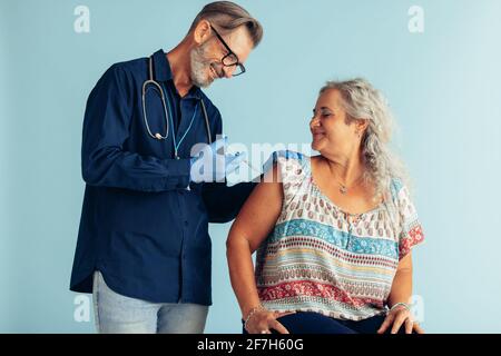 Ältere Frau, die von einem Arzt eine Grippeimpfung erhalten hat. Arzt, der älteren Frauen auf blauem Hintergrund eine Impfung verabreicht. Stockfoto