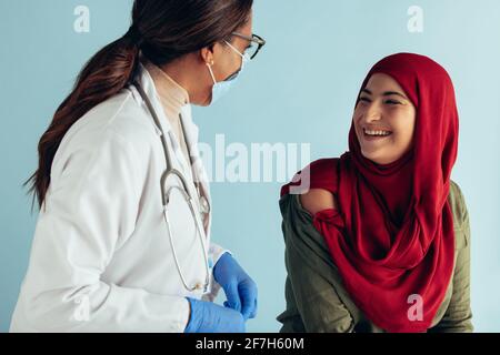 Lächelnde Frau im Hijab im Gespräch mit Ärztin in Gesichtsmaske. Frau, die von einem Arzt impfen wird. Stockfoto