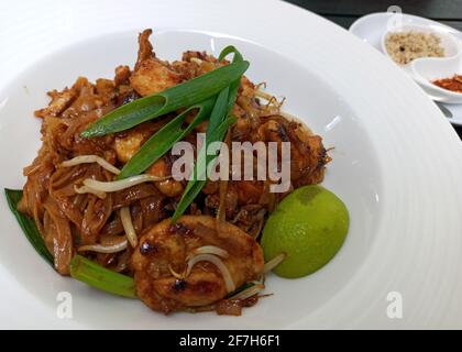 Pad thai Nudeln mit gebratenem Huhn, gemischte grüne Blätter, gehackte Erdnüsse, Schalotten & Limettenkeil auf weißem Teller vor Holzhintergrund serviert heiß wi Stockfoto