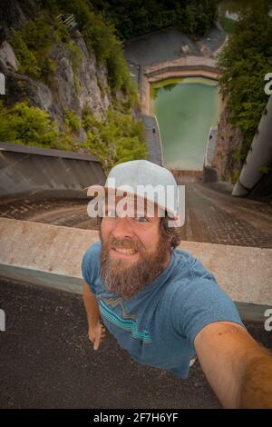 Hipster-Mann hat Angst, ein Selfie aus einem kleinen, aber steilen Staudamm für ein Wasserkraftwerk in Moste, Slowenien, zu machen. Blick auf den hidroelektrischen Damm von oben, siehe Stockfoto