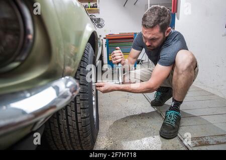 Junger Mann, der Räder oder Reifen aus einem alten Oldtimer aus den 60er oder 70er Jahren in seiner Garage abzieht. Werkzeuge sind überall zu sehen. Stockfoto