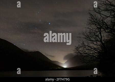 Brotherswater in einer bewölkten Nacht mit orion, der durch die sticht Wolken und ein Autokopf leuchten Nebel auf & Silhouette eines Baumes & die Umgebung Stockfoto