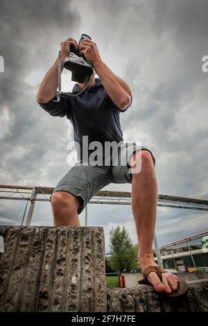 Mann in Flip-Flops, der ein Bild einer Fabrik mit einer analogen Vintage-Kamera aus Froschsicht und mit Wolken im Hintergrund fotografiert. Stockfoto