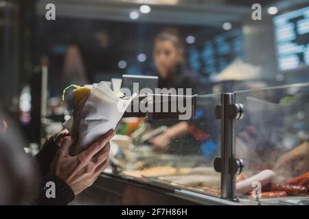 Person, die einen frischen Kebab oder Gyros in den Händen vor einem Fast-Food-Händler oder -Stand hält. Kebab in weißes Papier eingewickelt. Stockfoto