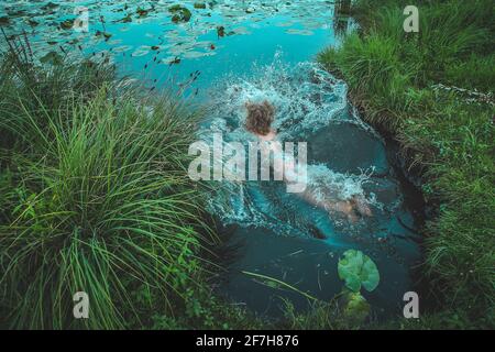 Eine Frau, die in einem Teich oder See springt, umgeben von großem Gras und Blättern. Der Rücken einer Frau und ihre lockigen Haare werden gesehen, wie sie ins Wasser gelangen. Stockfoto