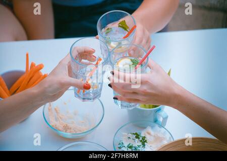 Hände von drei Frauen, die von oben betrachtet wurden, sahen Jubel oder Toasten mit Gläsern Gin Tonic Drink. Andere gesunde Lebensmittel sind um zu sehen, wie Dips und c Stockfoto