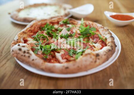 Nahaufnahme einer leckeren Pizza mit Schinken, Rucola und einer weiteren Pizza mit verschiedenen Käsesorten und roter Sauce. Konzept von köstlichen Mittag-oder Abendessen mit leckeren Pizza auf weißem Teller im Café. Stockfoto