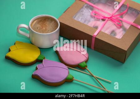 Lebkuchen in Form von Gelb. Rosa und lila Tulpen mit einer weißen Kaffeetasse und einer festlichen Box auf türkisfarbenem Hintergrund. Festliche Speisen im Hintergrund. Stockfoto