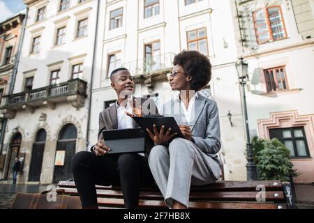 Hübscher Mann im Anzug, der auf einem digitalen Tablet arbeitet, während eine attraktive Frau auf dem Handy spricht und auf der Zwischenablage schreibt. Zwei afrikanische Kollegen lösen Arbeitsprobleme außerhalb des Büros. Stockfoto