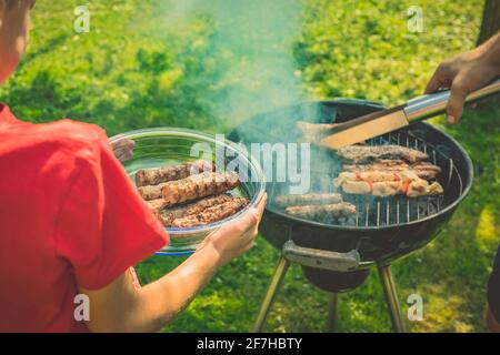 Unbekannter Junge in einem roten Hemd nimmt Fleisch vom Grill, während er eine Glasplatte oder Schüssel mit Fertigfleisch hält. Runder Grill in Rauch mit Cevapcici auf Stockfoto