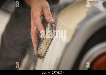 Eine Hand eines Mannes wird gesehen, wie sie einen alten Oldtimer mit einem Holzblock und braunem Sandkorn-Papier schleift. Manuelles Trockenschleifen eines Autos in der Restaurierung, proc Stockfoto