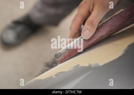 Eine Hand eines Mannes wird gesehen, wie sie einen alten Oldtimer mit einem Holzblock und braunem Sandkorn-Papier schleift. Manuelles Trockenschleifen eines Autos in der Restaurierung, proc Stockfoto