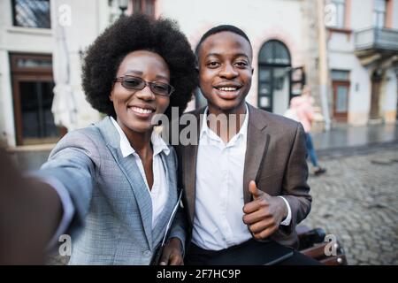 Lächelnder afrikanischer Mann und Frau im Business-Anzug stehen auf der Straße der Stadt zusammen und machen Selfie auf einem modernen Smartphone. Glückliche Partner im Freien. Stockfoto