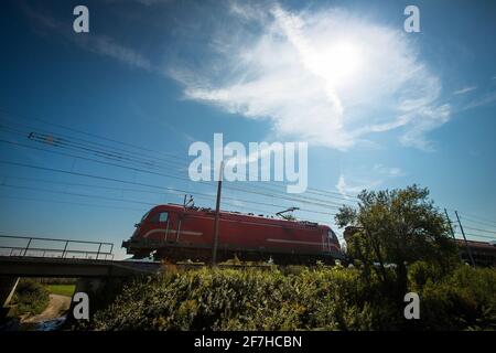 Die moderne europäische Lokomotive in roter Farbe schleppt an einem sonnigen Tag einen Güterzug über einen Damm. Stockfoto