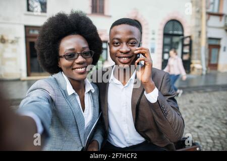 Lächelnder afrikanischer Mann und Frau im Business-Anzug stehen auf der Straße der Stadt zusammen und machen Selfie auf einem modernen Smartphone. Glückliche Partner im Freien. Stockfoto