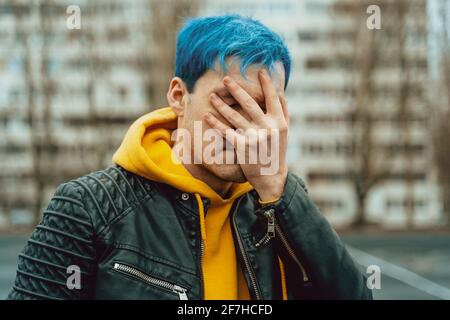 Porträt eines jungen Mannes, der sein Gesicht mit der Hand auf dem Hintergrund eines Hochhauses schließt. Hübscher Kerl mit blauen Haaren posiert im Frühling auf der Stadtstraße Stockfoto