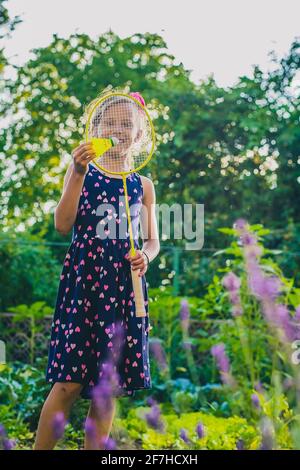 Junge kaukasische Mädchen oder Kind spielt Badminton-Spiel in einem üppigen grünen Garten. Kind hält einen Badmintonschläger vor dem Gesicht Stockfoto