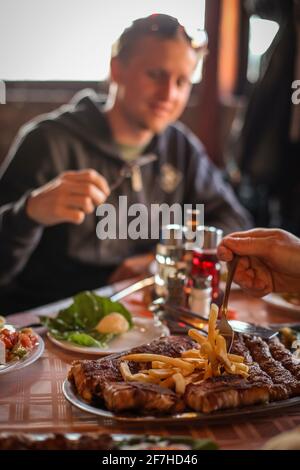 Der Mann, der glücklich hinter dem typisch serbischen Gericht sitzt, das aus Cevapcici, Pommes und Kajmak besteht. Eine Hand nimmt das Essen mit einer Gabel auf. Stockfoto